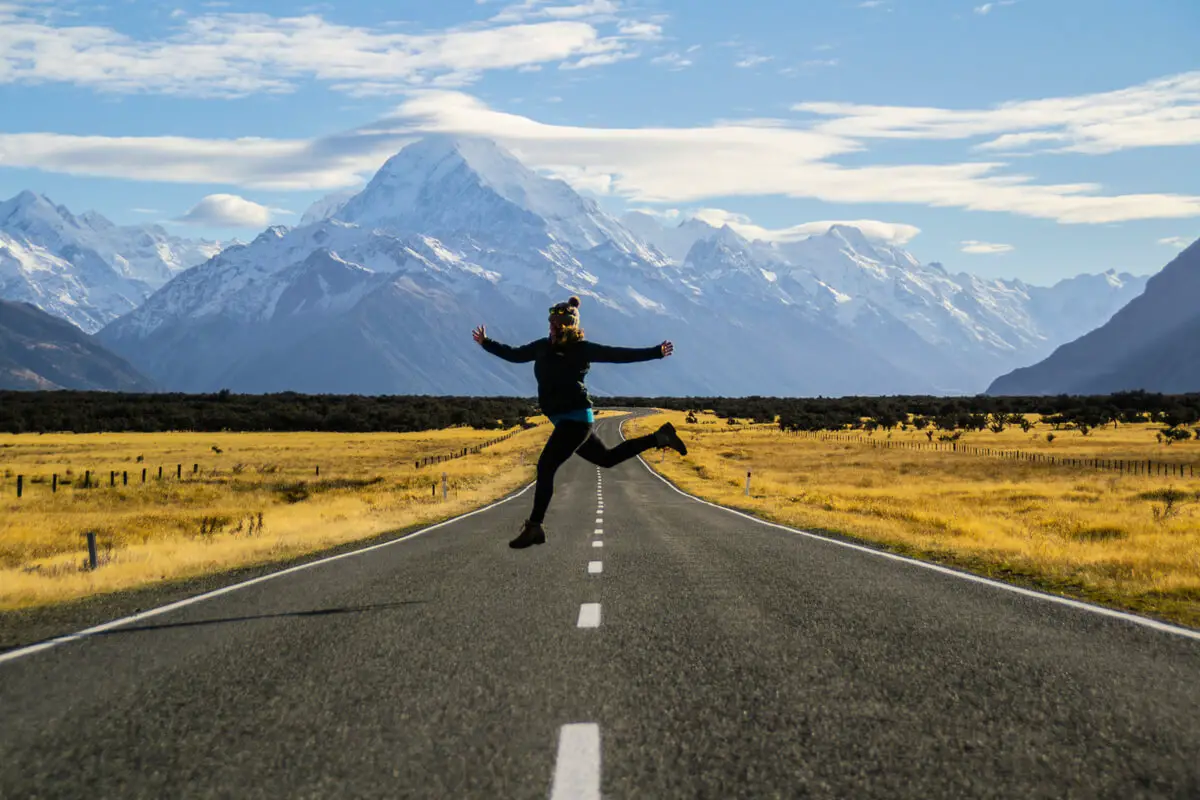 A girl jumps in the middle of a road with the snow capped mountains in the distance in new zealand