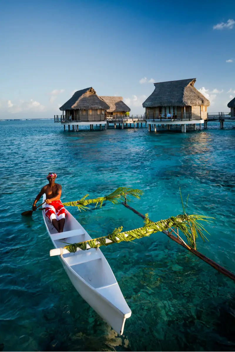 A man in a canoe rows past thatched roof houses over clear blue water in french polynesia
